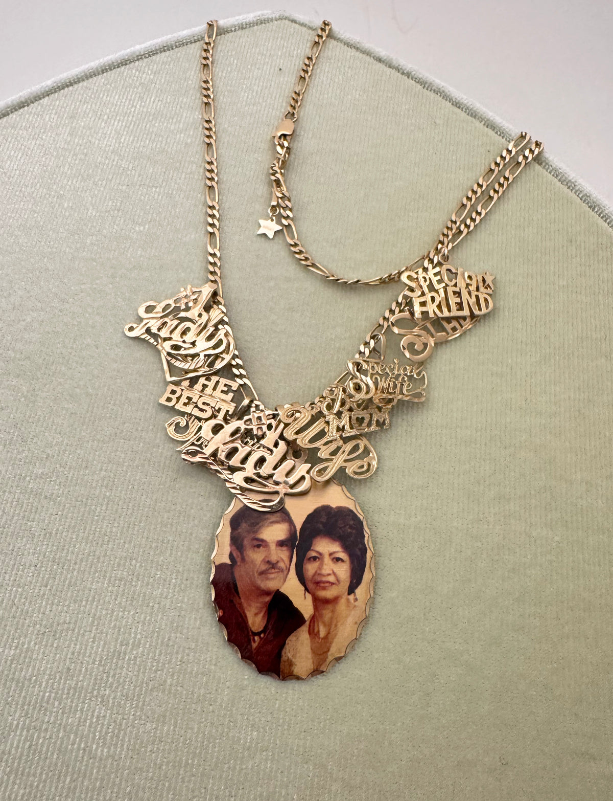 A 14K gold  personalized necklace displayed on a light sage green textured stand. The necklace features multiple script nameplate charms spelling out '#1 lady' and a teardrop-shaped pendant containing a custom photo of a couple.