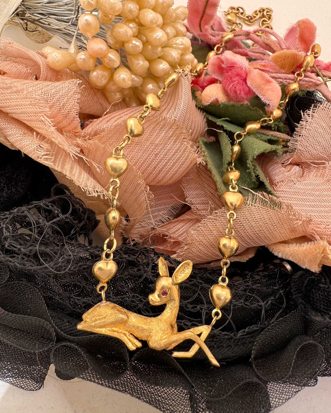 A gold-toned necklace featuring a deer pendant and heart-shaped beads, styled on a black lace surface with pink fabric and faux flowers in the background.