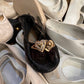 A gold heart-shaped ring with clear diamonds laying on a black velvet shoe, surrounded by a pile of off-white and beige shoes.