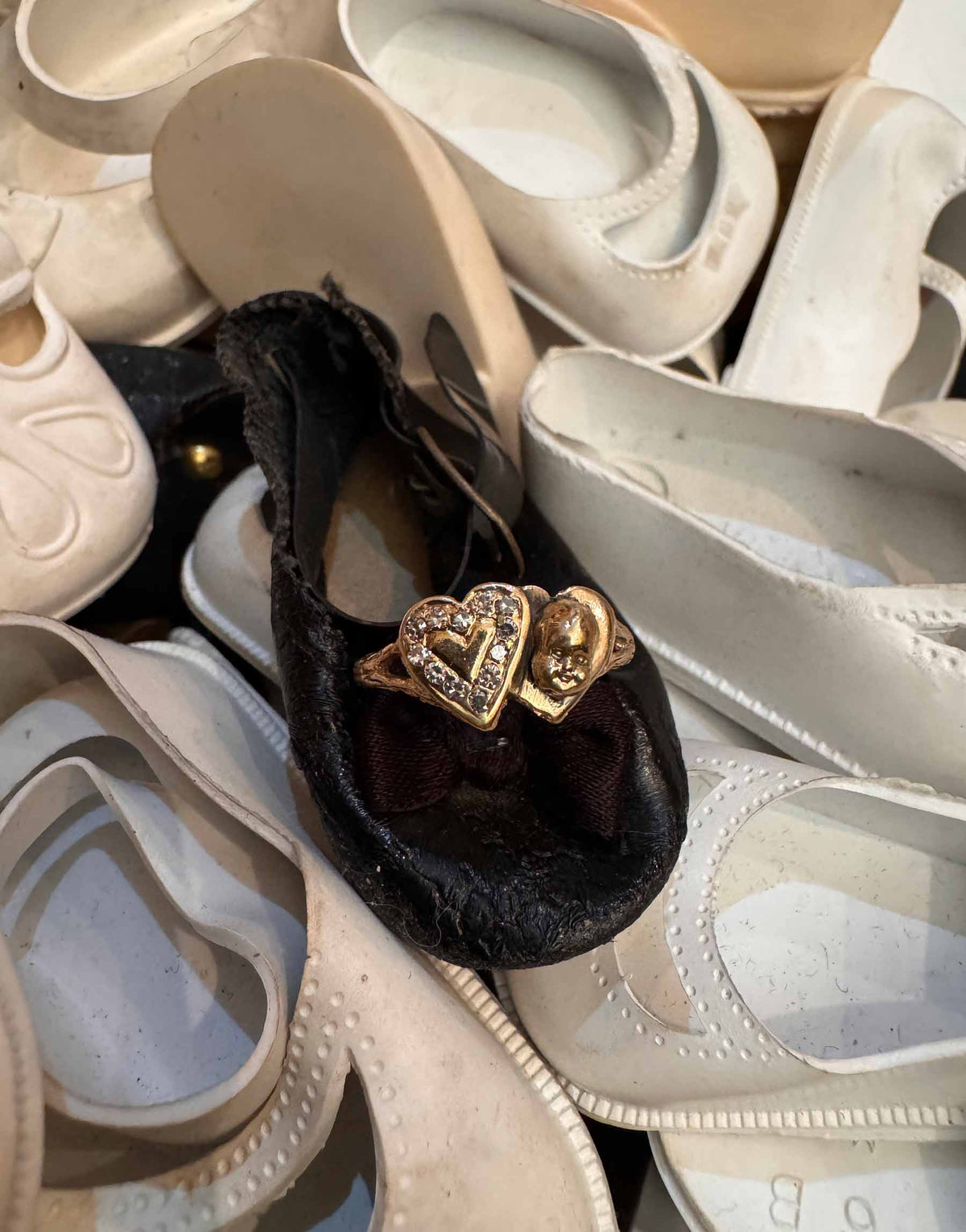 A gold heart-shaped ring with clear diamonds laying on a black velvet shoe, surrounded by a pile of off-white and beige shoes.