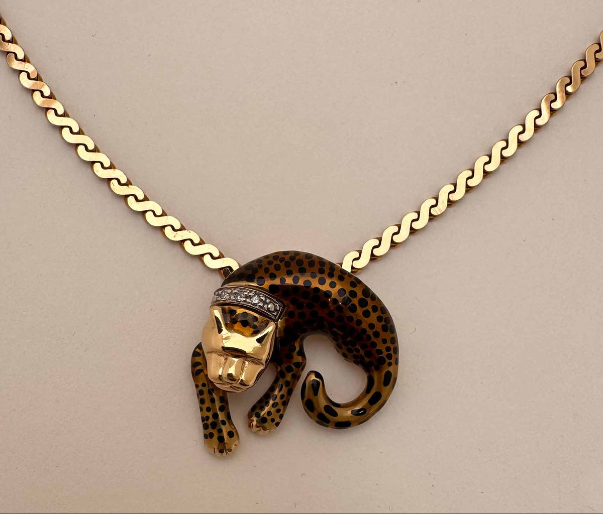A gold chain necklace featuring a leopard pendant with a gold and black spotted pattern and a small diamond crown, displayed on a neutral beige background.
