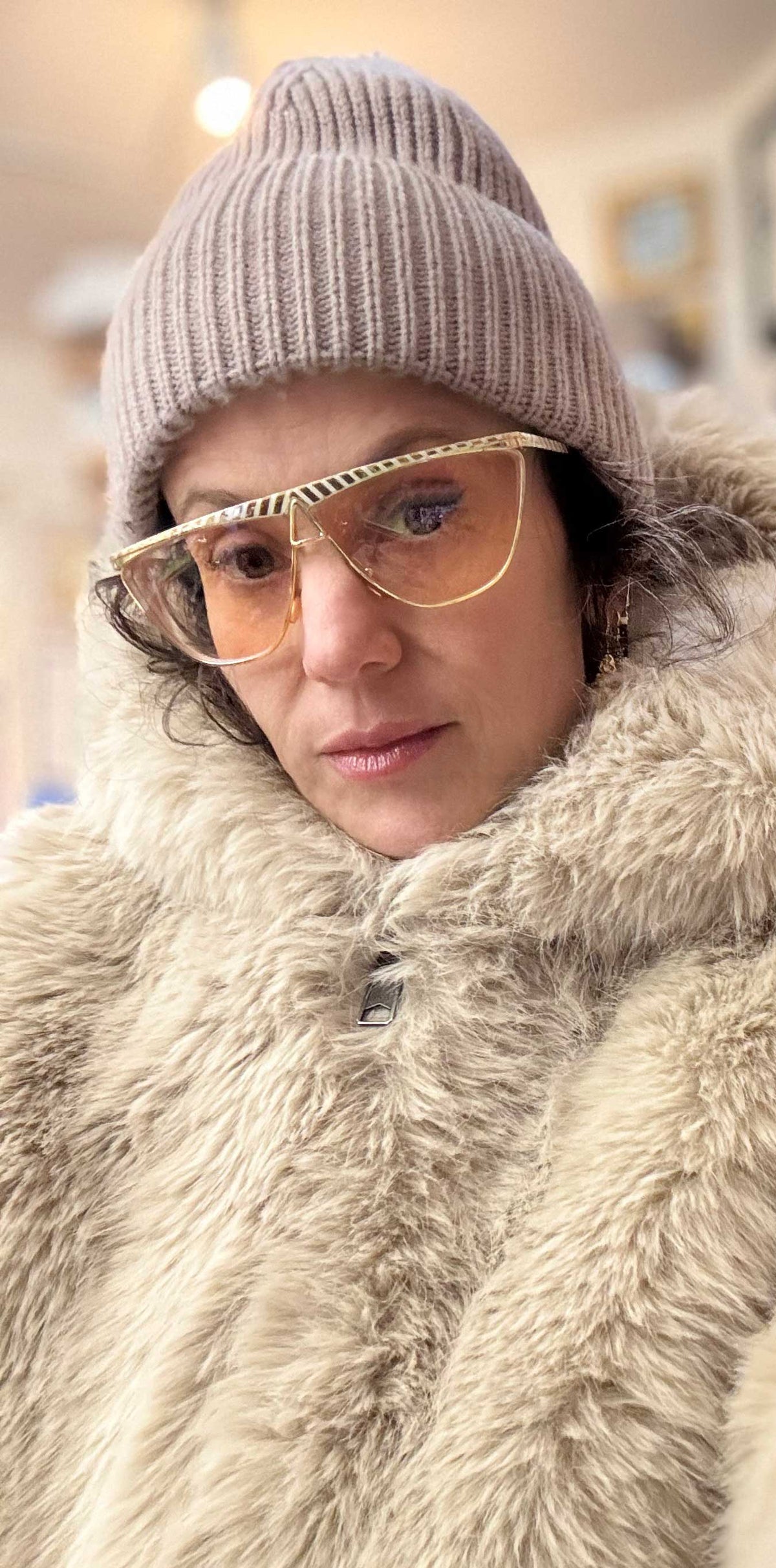 Karen yost, ownder and maker of Anomaly RElic in New York City wearing a thick beige faux fur coat with a high fluffy collar, a ribbed taupe beanie, and gold-framed glasses with amber lenses, photographed in a softly blurred indoor setting.