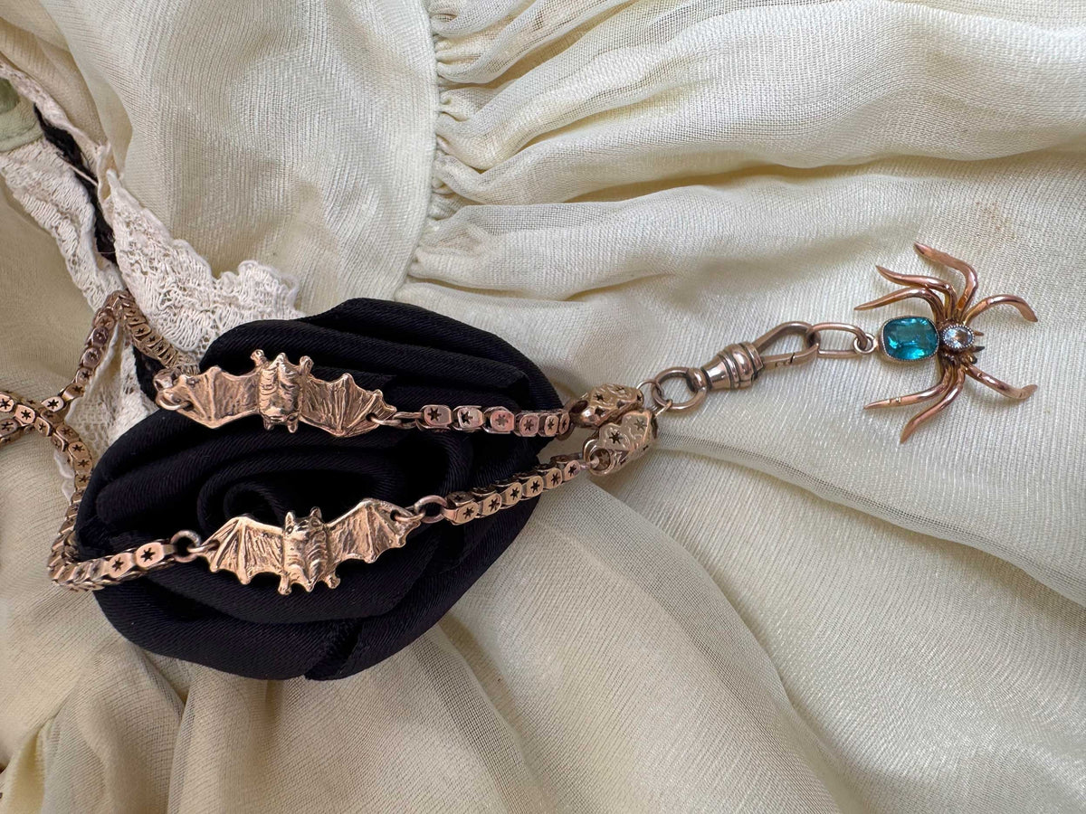 A gothic-style necklace featuring 14K gold bat charms on an antique pierced star chain, draped over a black fabric rose and cream-colored fabric
